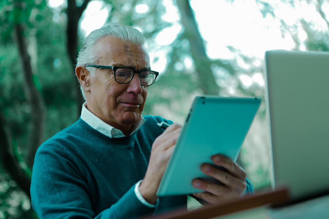 old businessman with an iPad on hand and a laptop on a bench table